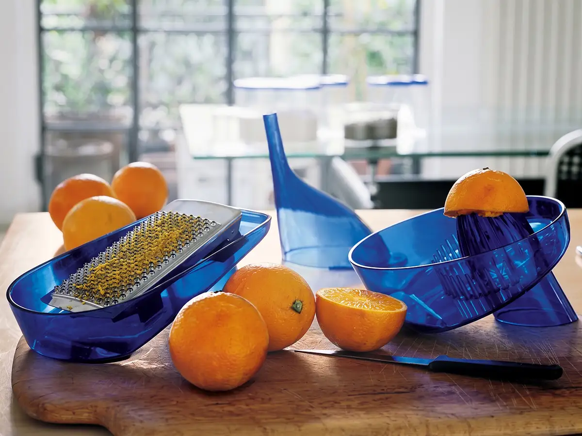 Grattugia, imbuto e spremiagrumi della stessa serie in plastica blu trasparente su un tagliere, con arance