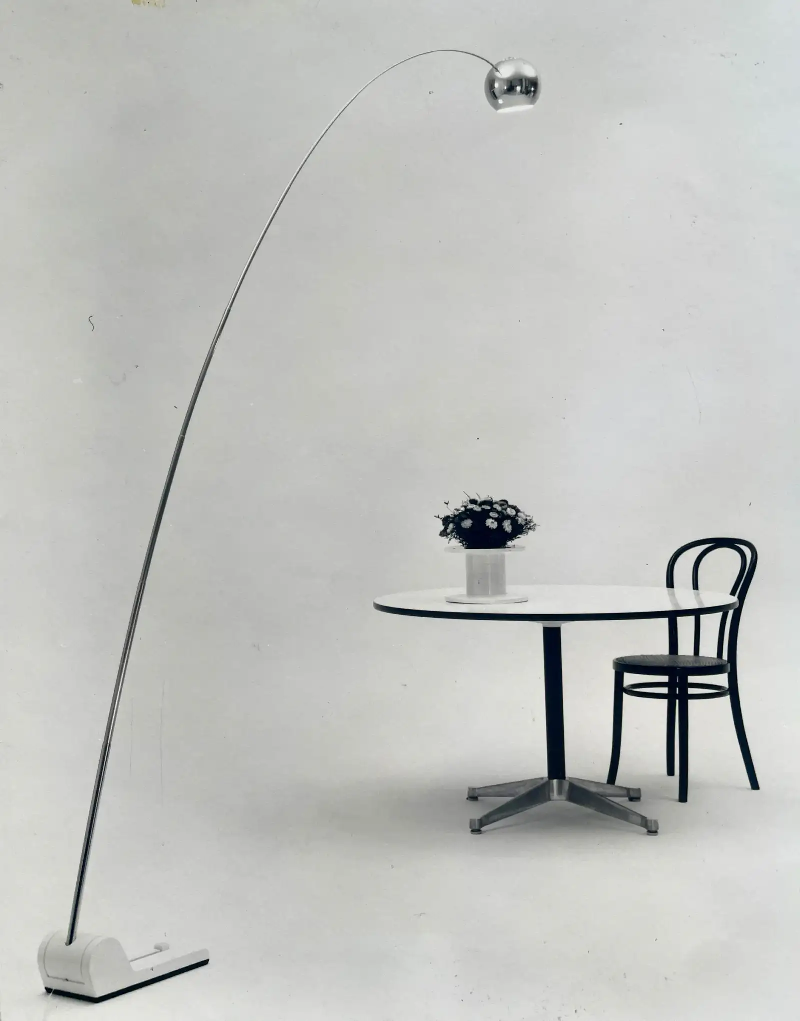 Foto in bianco e nero di una lampada da terra con lungo stelo ad arco sopra un tavolo tondo con una sedia, Reggiani
