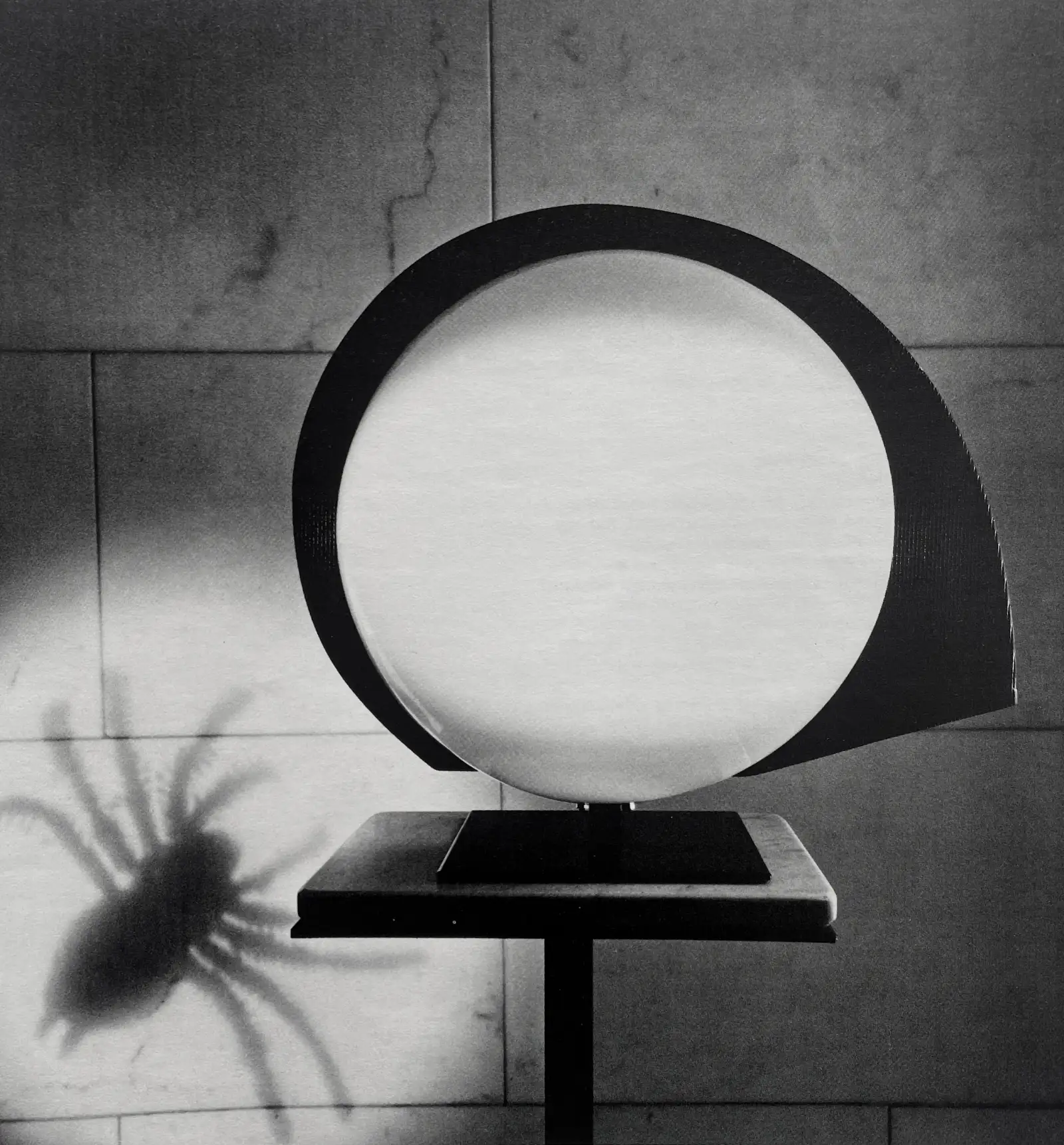 Lampada da tavolo Polifemo con schermo circolare, con l’ombra di un ragno sulla parete; foto in bianco e nero, Arteluce Flos