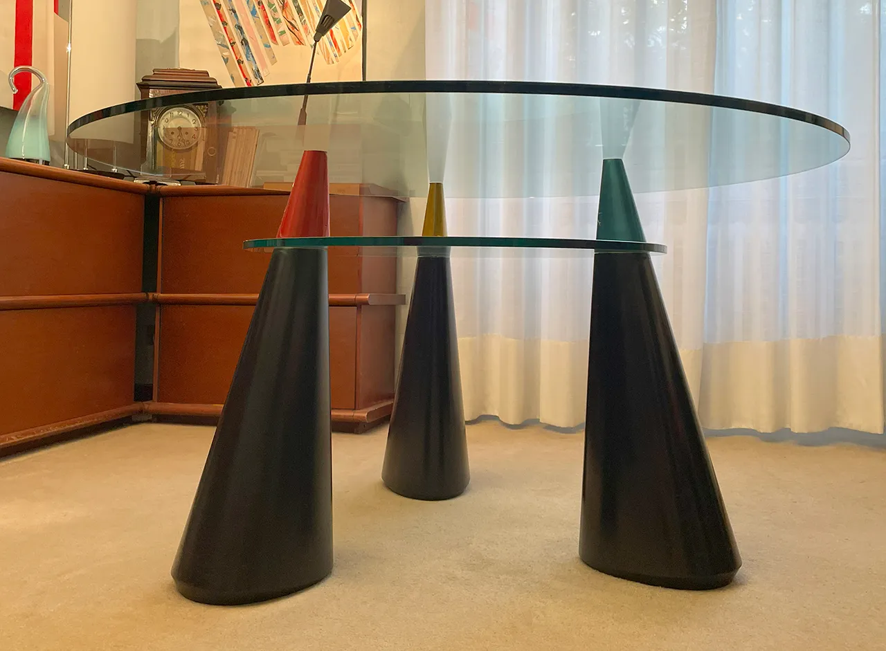 Il prototipo del tavolo visto all’altezza del piano: i due piani in vetro, i coni distanziali colorati e le gambe scure, in una stanza con un mobile in legno e tende chiare