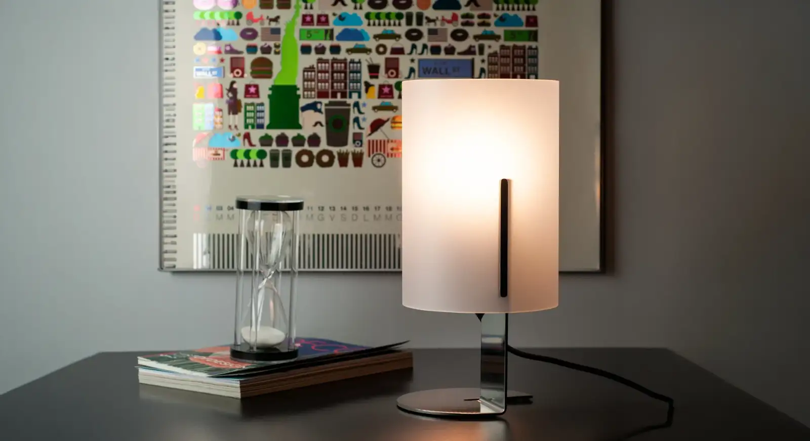Lampada da tavolo Quarter accesa su un mobile scuro, con schermo bianco curvo, Vesta