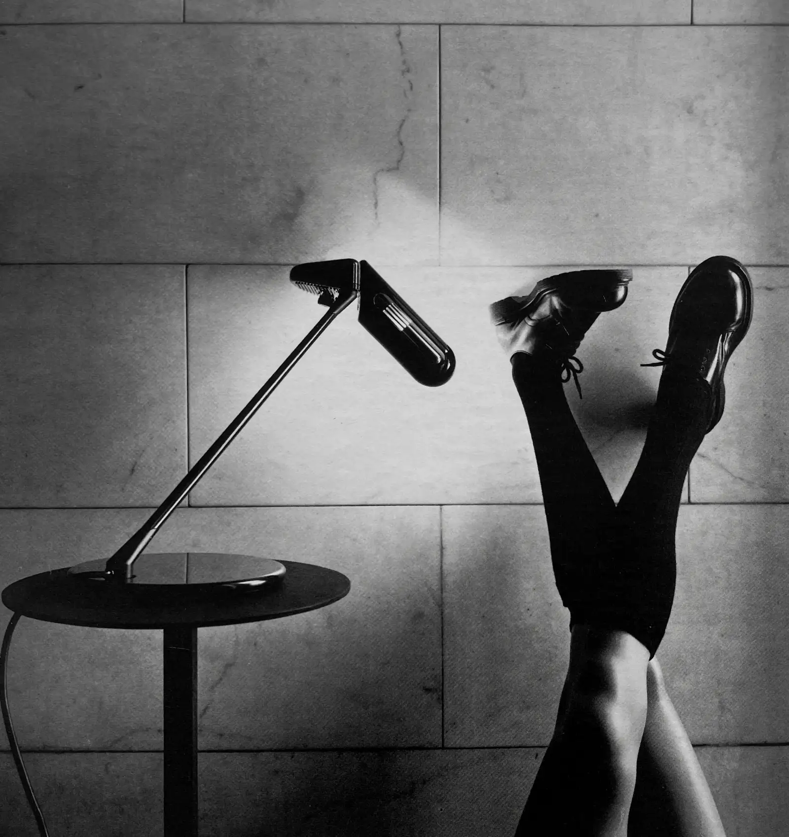 Lampada da tavolo alogena Ring su un tavolino, accanto a due gambe con scarpe col tacco; foto in bianco e nero, Arteluce Flos